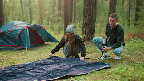 man pumps tent bag while wife checks air level smiling as he looks at her lovingly during outdoor camping trip surrounded by forest trees and tent