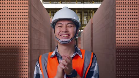 close up of asian male engineer with safety helmet smiling and clapping his hands while standing with red brick packed in stacks are stored