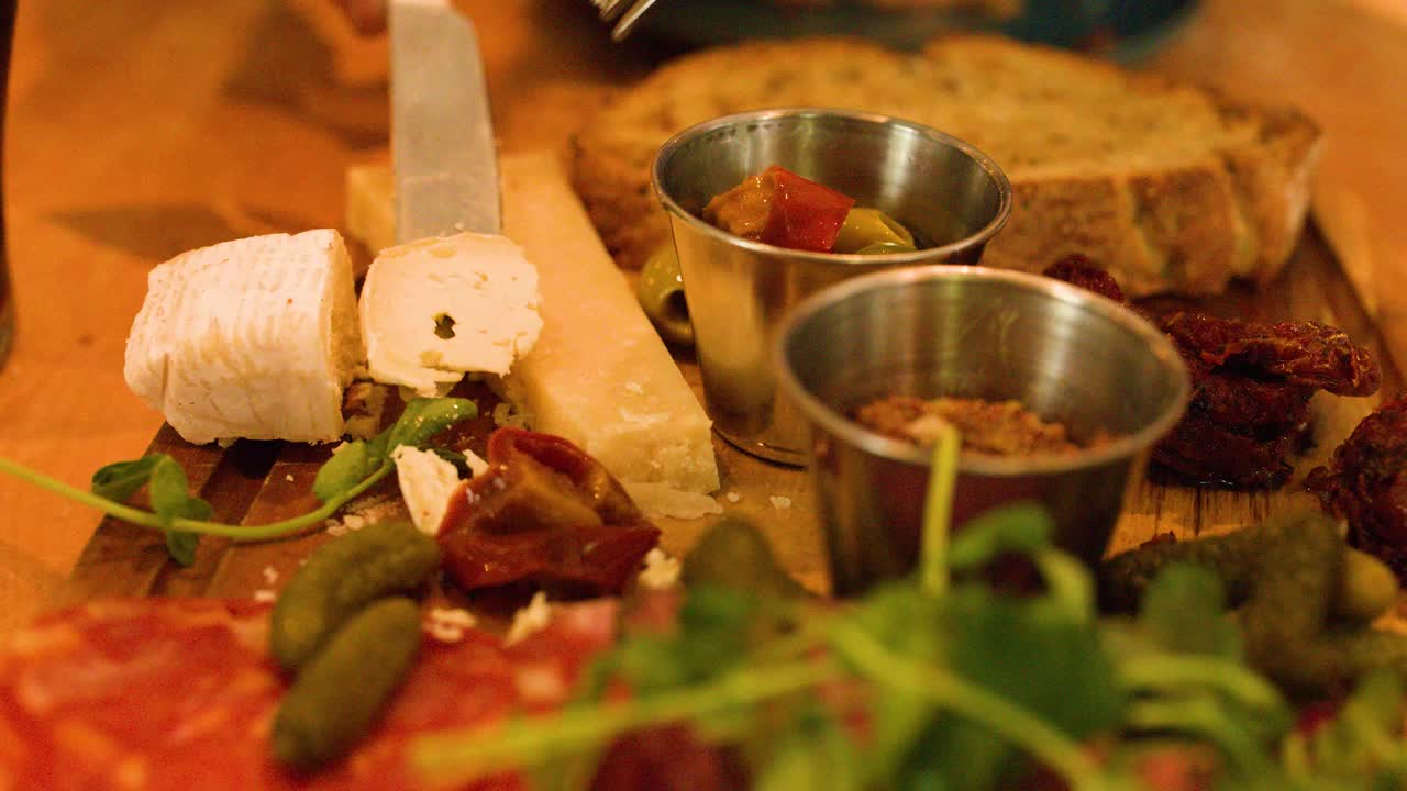 Hand slices soft cheese on charcuterie board with meats, pickles, bread. Warm, inviting lighting