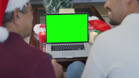 Padre-Birracial-Sonriente-Con-Hijo-Con-Sombreros-De-Papá-Noel-Haciendo-Videollamada-Navideña-Con-Computadora-Portátil,-Con-Pantalla-Verde