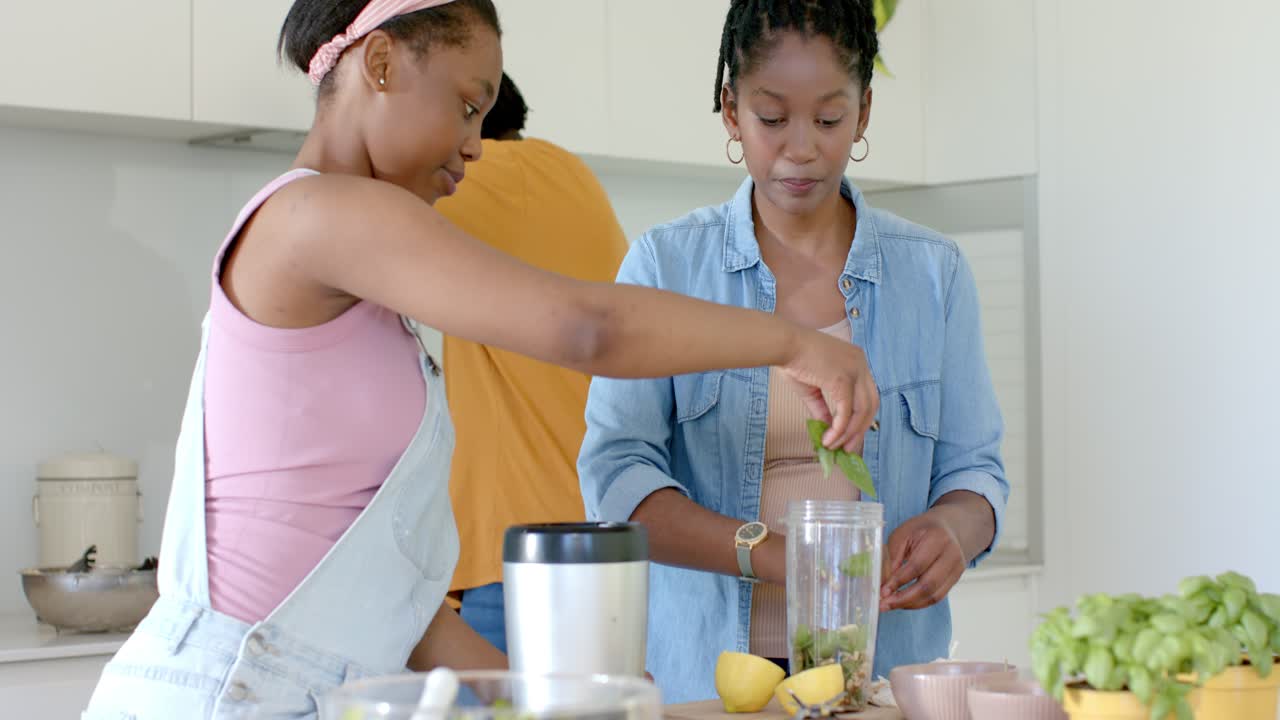 Afrikansk mor og datter laver mad ved køkkenbordet, hvor den unge rækker ud og tilføjer grøntsager til blenderkrukken.