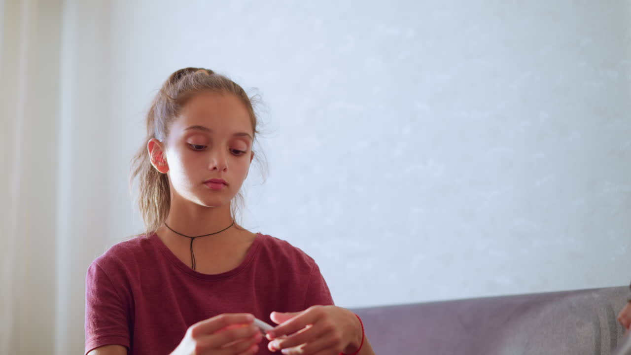 Young girl with ponytail and maroon shirt checks thermometer reading while sitting beside sick person resting off-frame showing care concern and responsibility in calm indoor setting