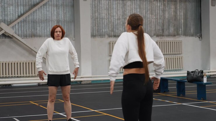 Campo da badminton indoor, due donne che si preparano per gli esercizi, figura dell'allenatore che si avvicina alla giocatrice, scambio di istruzioni vicino alle linee segnate e alle panchine, atmosfera calma e concentrata con riscaldamento pratico.