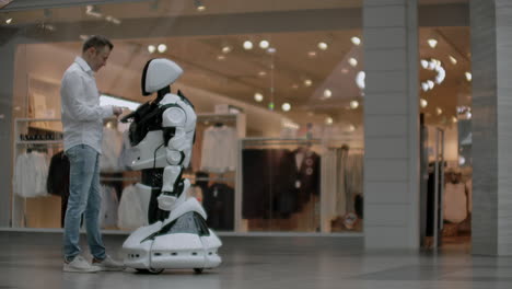 Un-Hombre-Con-Camisa-Se-Comunica-Con-Un-Robot-Blanco-Haciendo-Preguntas-Y-Presionando-La-Pantalla-Con-Los-Dedos