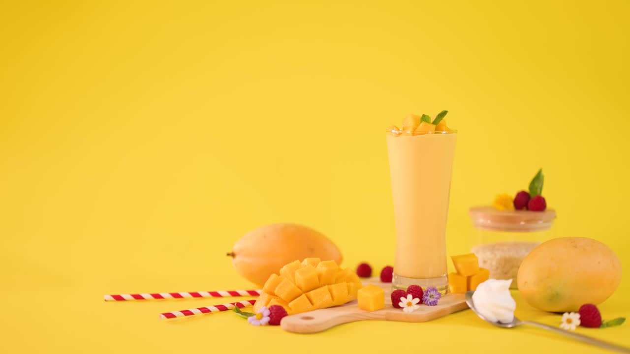 Una mano umana decora un alto frullato di mango tra frutta fresca su uno sfondo giallo brillante da studio
