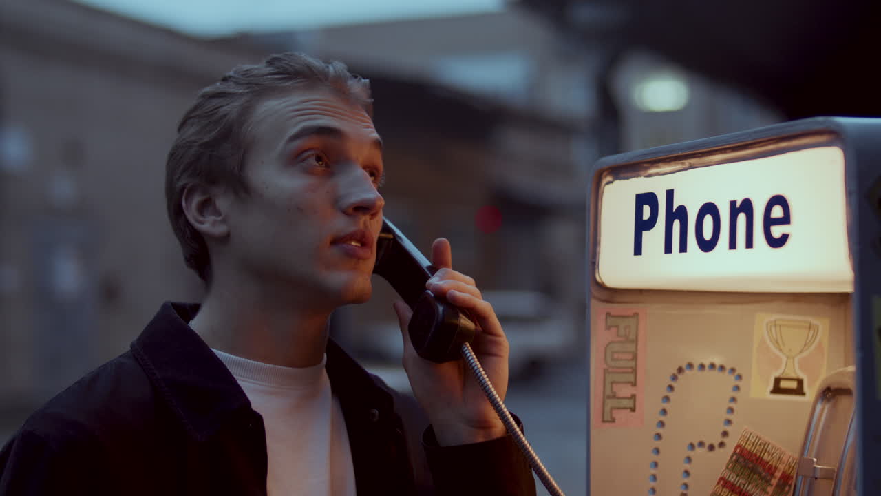 Young Man Talking on Pay Phone Outdoors on City Street in Evening