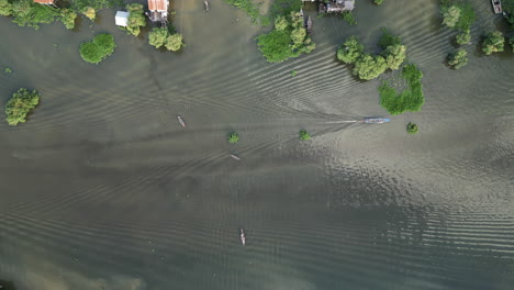 drone view of a boat moving through tonlé sap waters in cambodia. ripples spread across the muddy river as small houses and trees line the banks