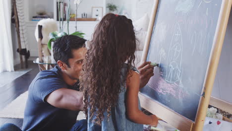 Father-sitting-on-the-floor-at-home-drawing-on-a-blackboard-with-his-young-daughter,-close-up