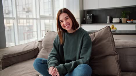 portrait of a happy brunette girl in a dark green sweater and jeans who is sitting on a modern brown sofa in a studio apartment