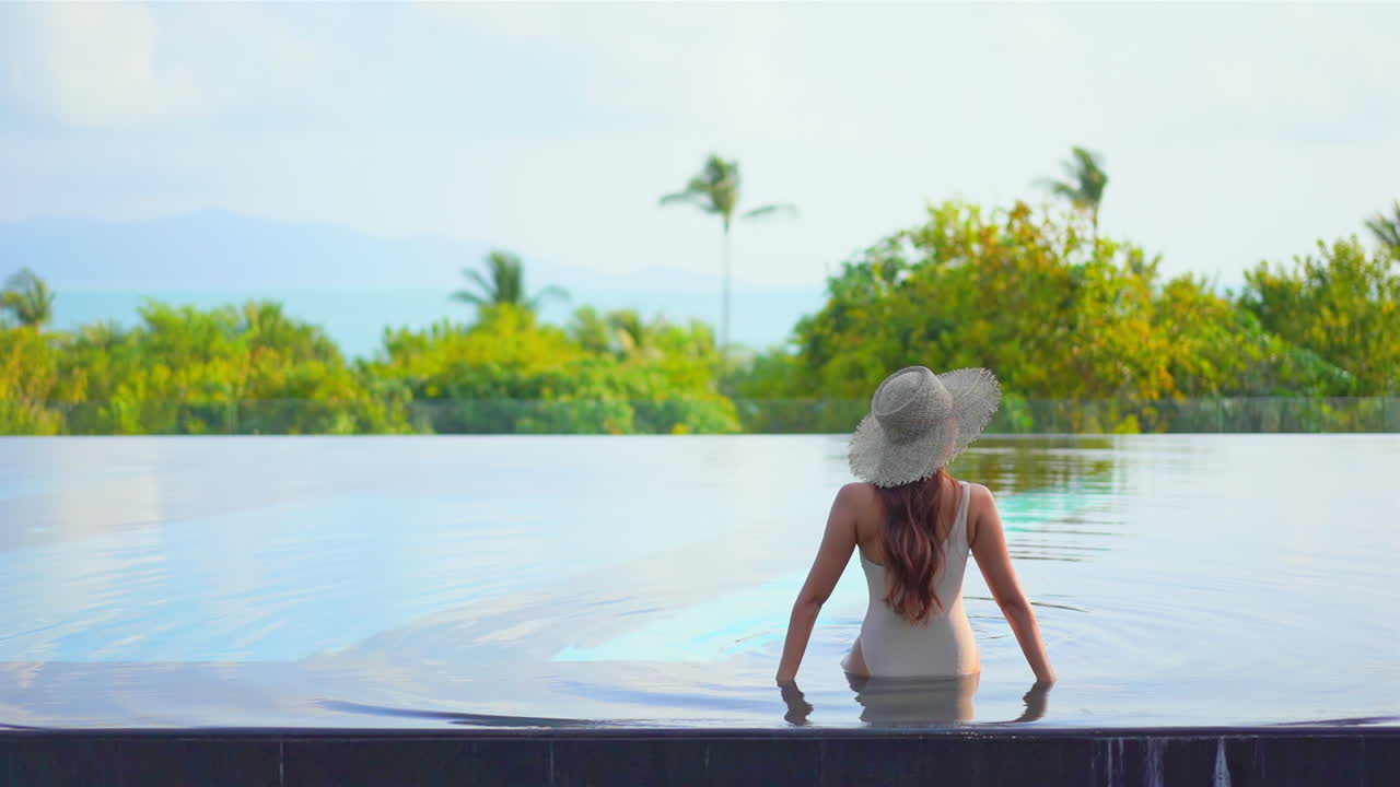 Model im Badeanzug und mit Hut sitzt am Rande eines Infinity-Pools und stützt sich auf die Arme vor dem Hintergrund tropischer Berge und grüner Bäume, Zeitlupen-Rückenansicht bei Tag