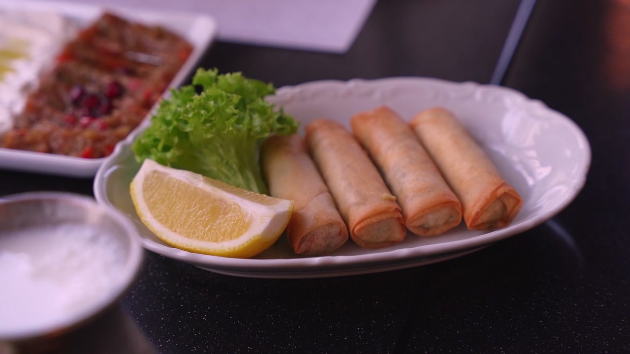 Close-up lumpia goreng disajikan dengan lemon dan selada di piring putih di restoran. Hidangan pembuka meze Mediterania otentik dan minuman ayran berada di latar belakang yang buram