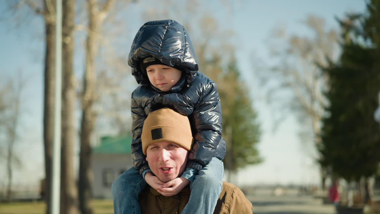 primer plano de un padre llevando a su hijo en sus hombros, sonriendo y hablando, con el niño vestido con una chaqueta negra brillante y capucha, con árboles borrosos y una persona con una camisa polo roja