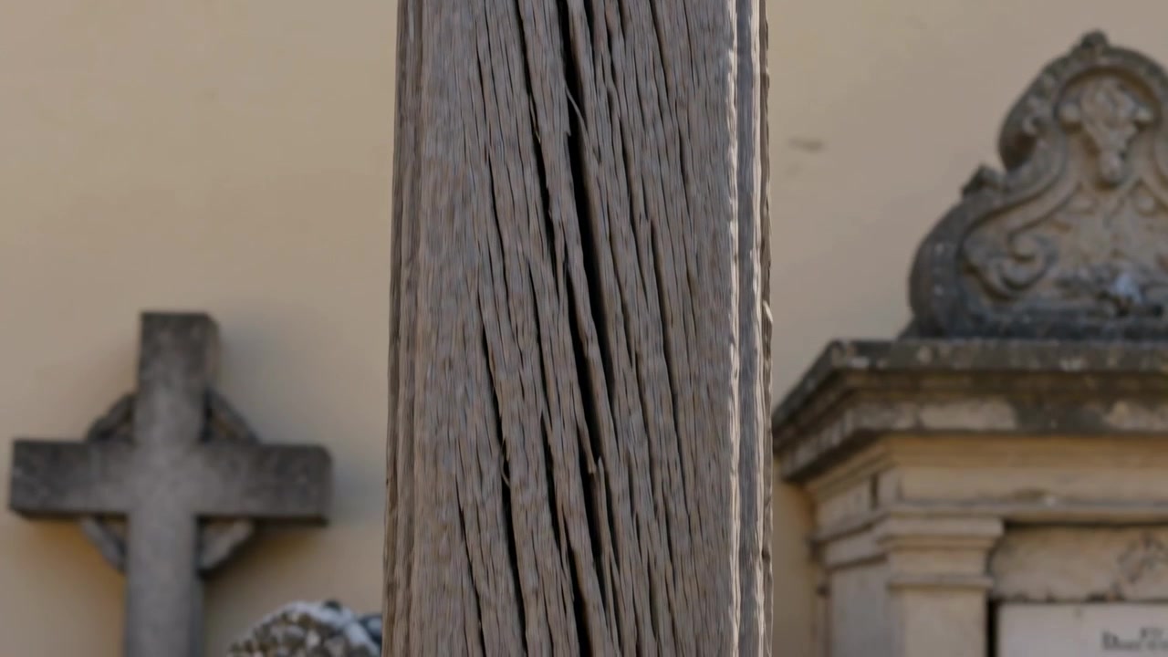 La telecamera si ritrae, mostrando un palo di legno invecchiato su un piedistallo di pietra in un cimitero con una croce di pietra.