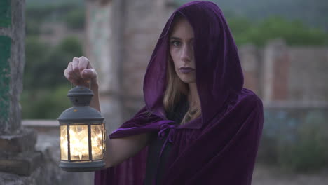 mysterious young woman wearing a cloak and a hood covering her head staring at the camera and holding an old lantern