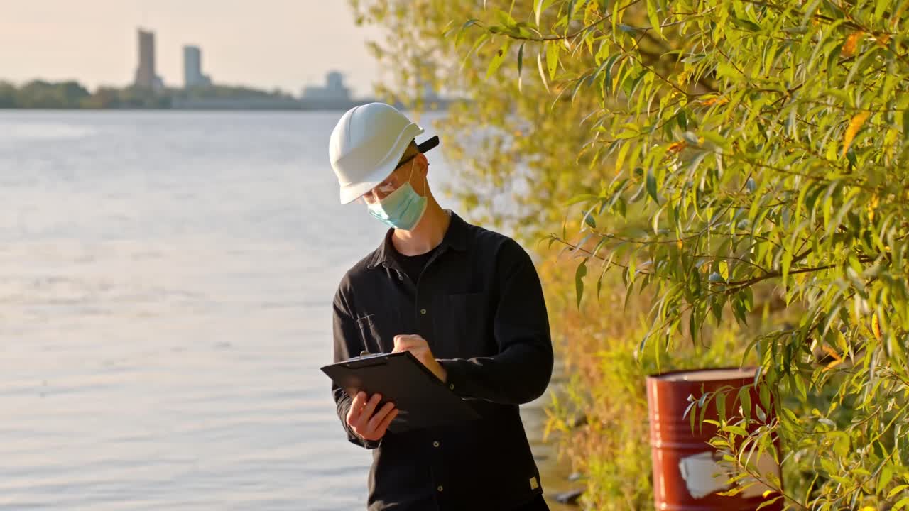 Scientist stands near lake wearing safety mask, taking notes on clipboard with oil drum behind