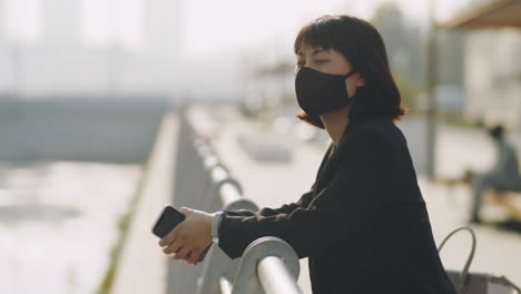 Asian-Businesswoman-in-Mask-Standing-on-Embankment-in-City