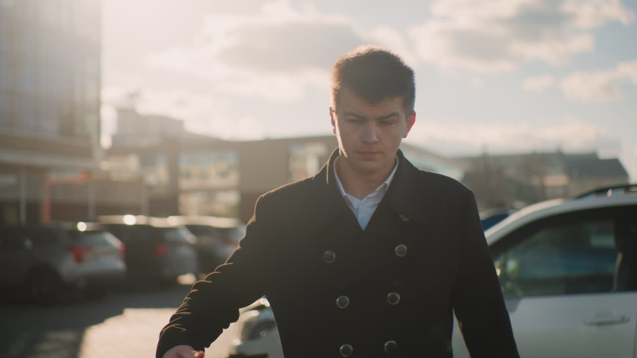 검은 코트를 입은 은행가는 차를 떠나 심각한 표정으로 주머니에서 전화를 꺼내서 전화에 응답합니다. 주차된 자동차와 현대적인 건물이 있는 활기찬 도시 지역에서