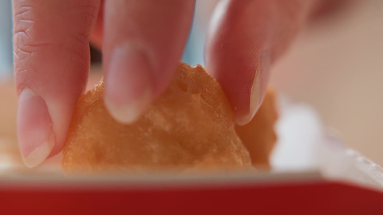 Low view showing two fingers pinching and lifting golden fried snack from red container edge, revealing crispy breadcrumb crust, slight surface indentation, warm directional light, soft shadow, and
