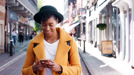 Mujer-Negra-Milenaria-Con-Un-Abrigo-Amarillo-Y-Un-Sombrero-Homburg-Usando-Su-Teléfono-Inteligente,-Parada-En-La-Calle-En-Un-Día-Soleado,-De-Cerca