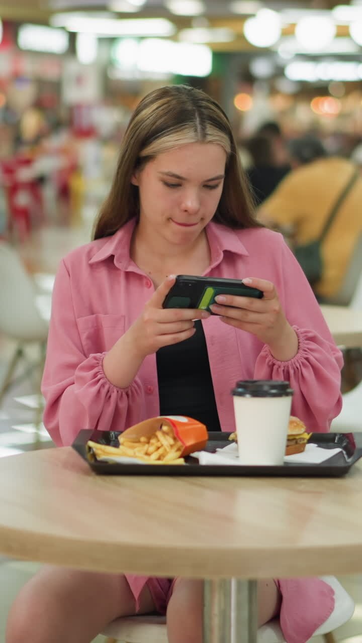 Lady in pink dress sits at table in busy restaurant, taking photo of her meal, she adjusts her position, standing slightly to capture the burger, fries, and coffee cup on black tray in front of her