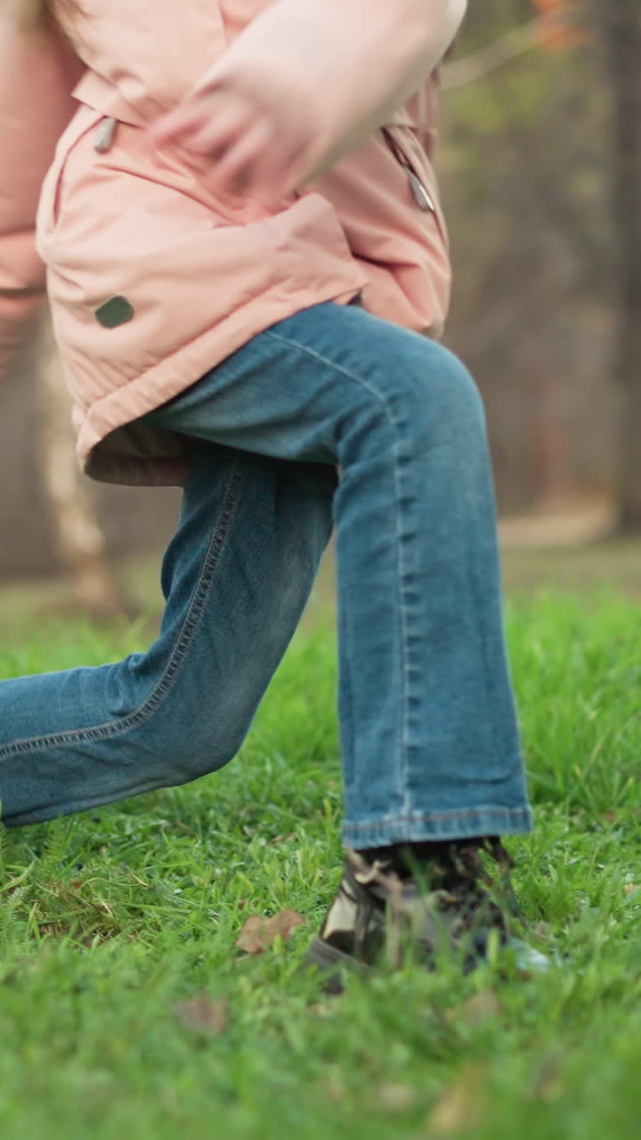 A blurred close-up shot features a girl and a man walking on lush green grass in a park. The girl, wearing a pink jacket, jeans, and black shoes, is in motion, with the background showing trees