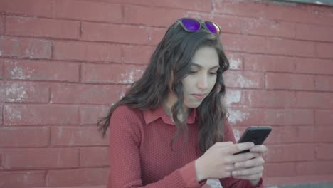 Chica-Enviando-Mensajes-De-Texto-Con-El-Teléfono-Inteligente-Con-Pared-De-Ladrillo-En-El-Fondo,-Pan