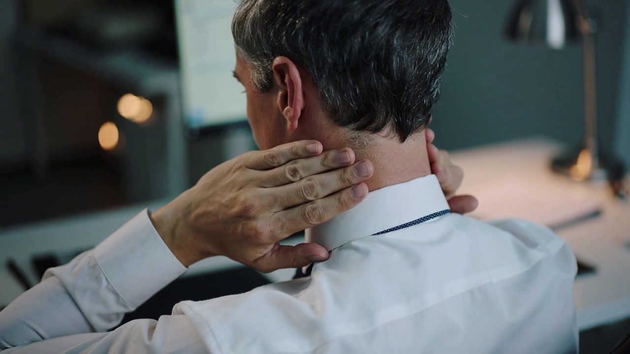In pausa al PC, uomo di mezza età in camicia bianca che si massaggia il collo con le mani alla scrivania per trovare sollievo