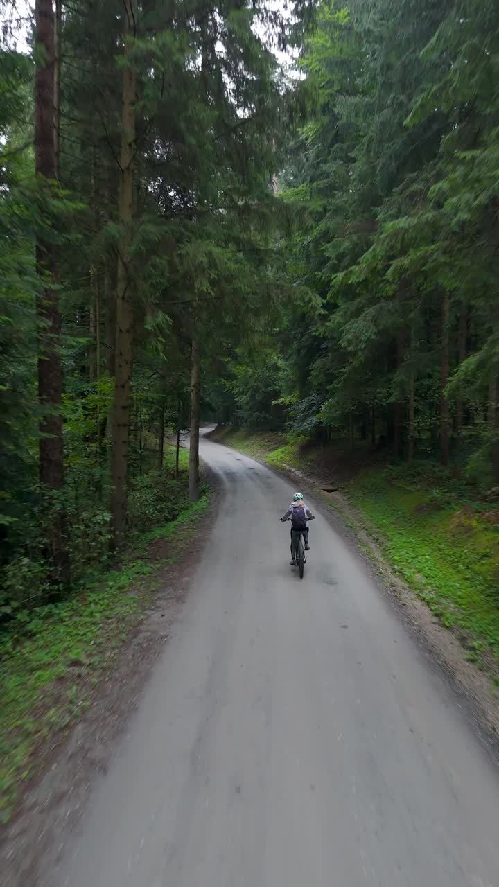 Vertical aerial tracking shot of woman riding e-bike on tarred forest path