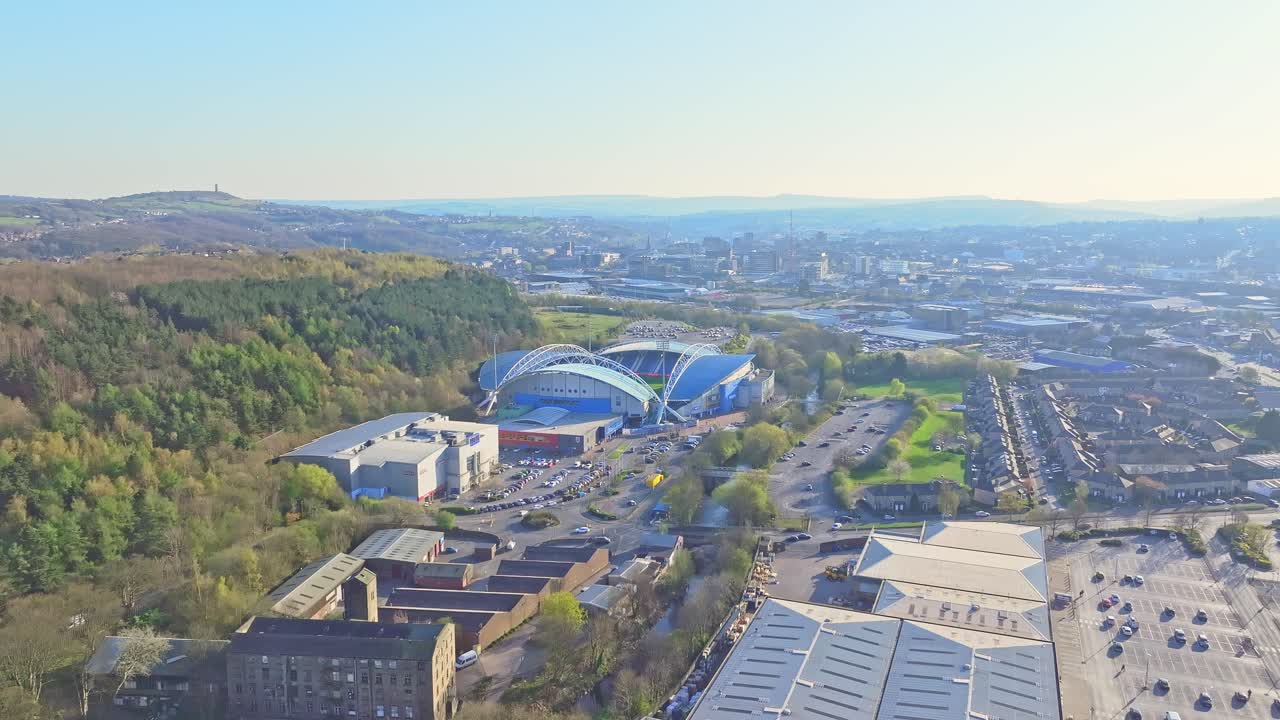 John Smith's Stadium in Huddersfield, adjacent River Colne, nearby woodland and parking areas, with Castle Hill and Victoria Tower visible on distant horizon under clear sky, drone static shot