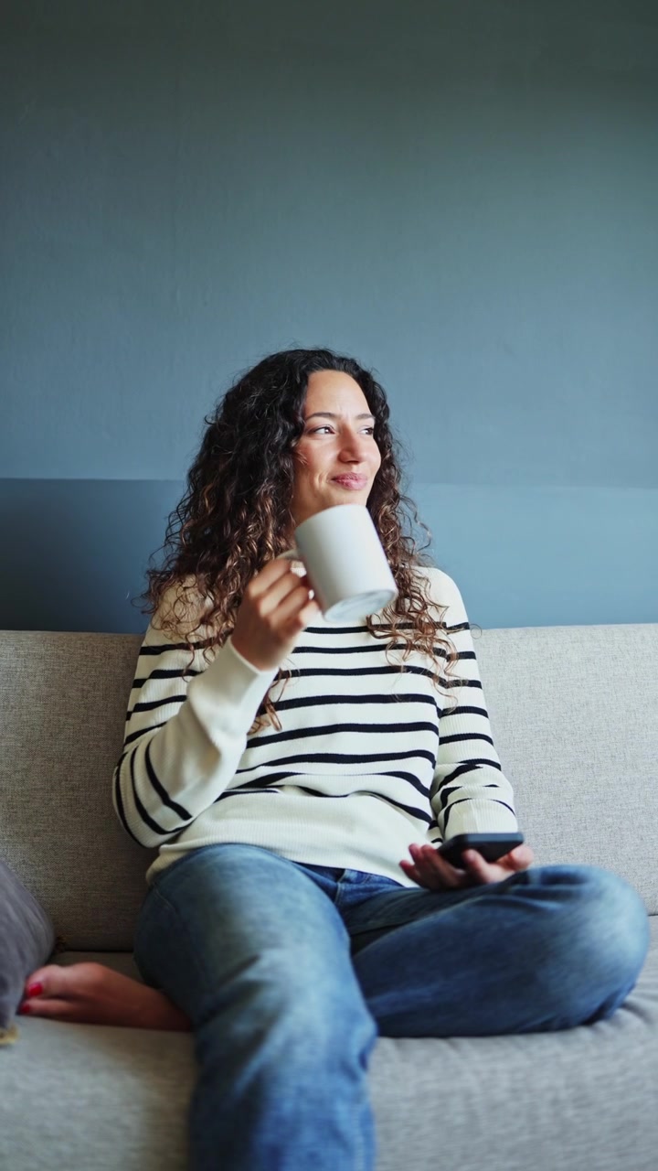 Giovane donna che si rilassa sul divano con smartphone e caffè.