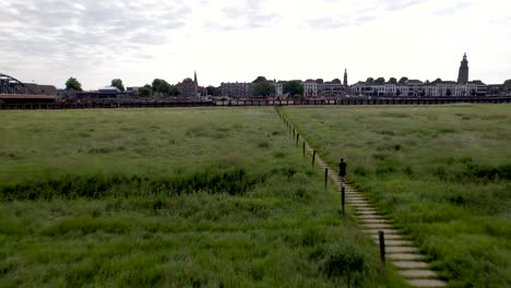 aerial following of male trail runner dutch floodplains with countenance of tower town zutphen along river ijssel behind