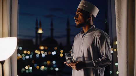 young muslim man wearing a taqiyah and a light blue shirt, holding a glass of water in his hand, looking out the window at the city lights at night, with a mosque in the background