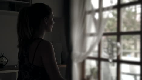 A-gentle-push-in-on-a-young-woman-sitting-in-a-yoga-pose-in-a-dark-room,-facing-the-light-of-a-nearby-window