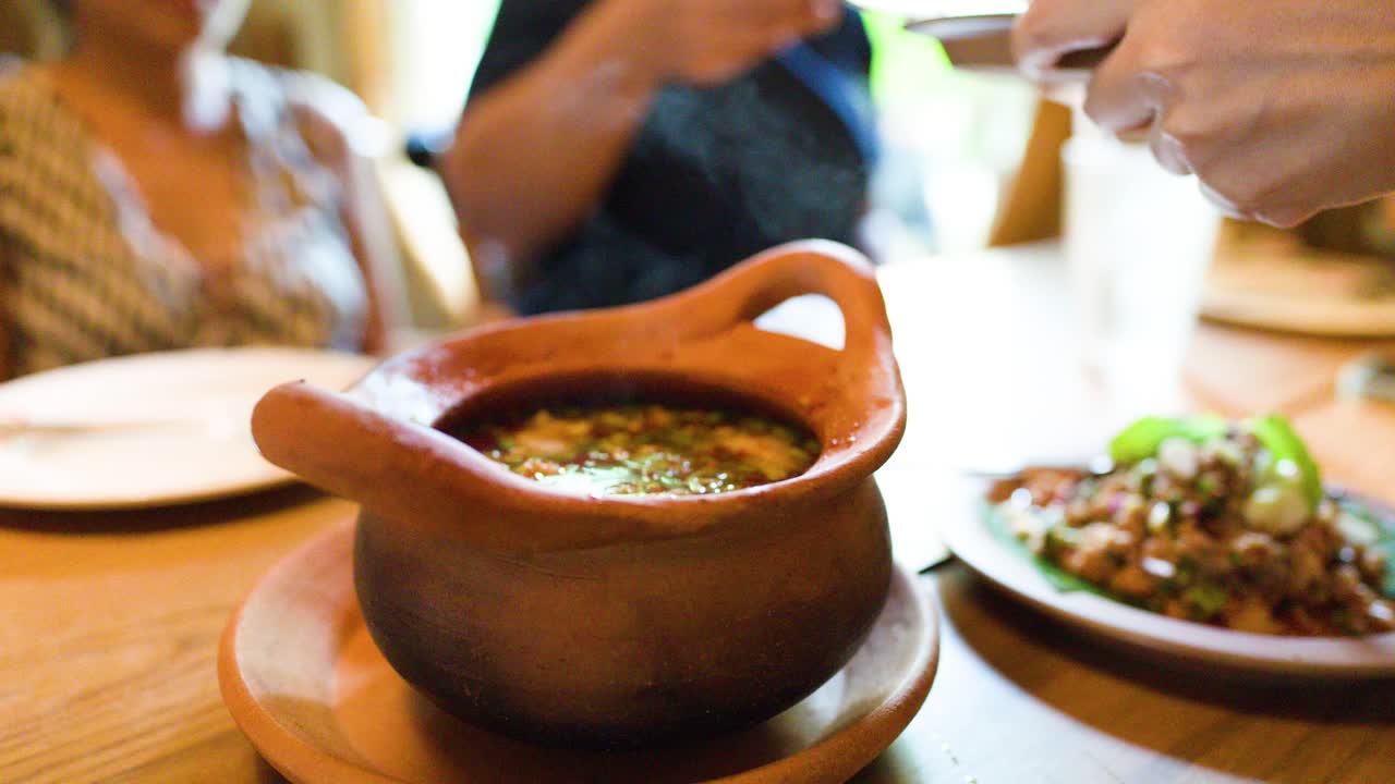 Diners reveal steaming herbal Thai soup in clay pot, sharing meal in warmly lit restaurant