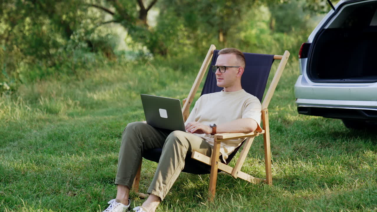 Freelancing male sits in a folding chair with laptop. Freelancer working remote in the nature.
