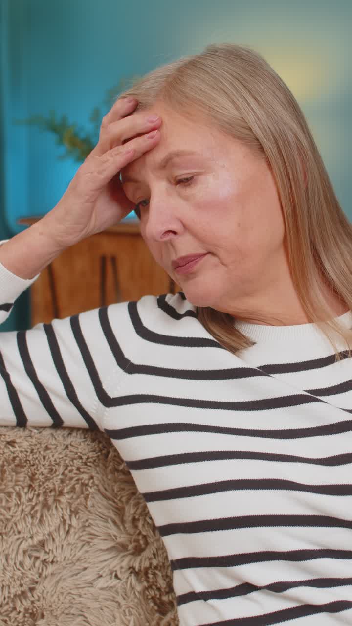 Mature woman placing hand on forehead on sofa showing frustration helplessness after bad day at home