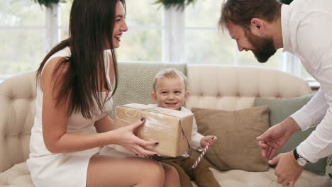 joven madre vestida de blanco recibiendo un regalo de navidad de su marido y besándolo. niño lindo rubio mirando su