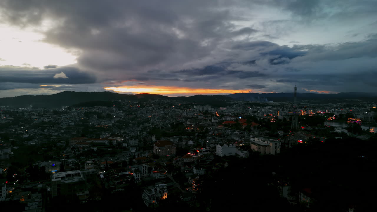 Vista aérea de un pueblo en la ladera de la montaña después del atardecer. Luces parpadeantes brillan en el valle bajo un cielo dramático con un horizonte dorado. Perfecto para viajes evocadores y tomas de establecimiento cinematográficas.