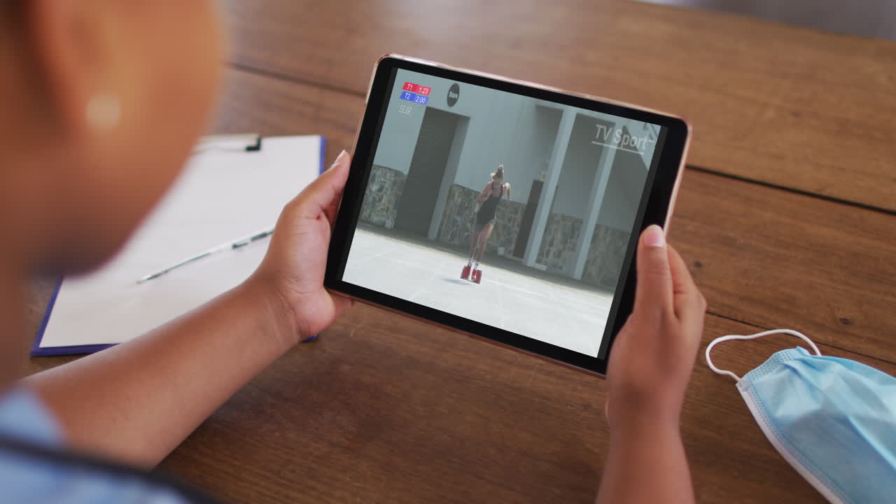 compuesto de una mujer sentada en una mesa con una máscara facial, viendo un evento de atletismo en la tableta