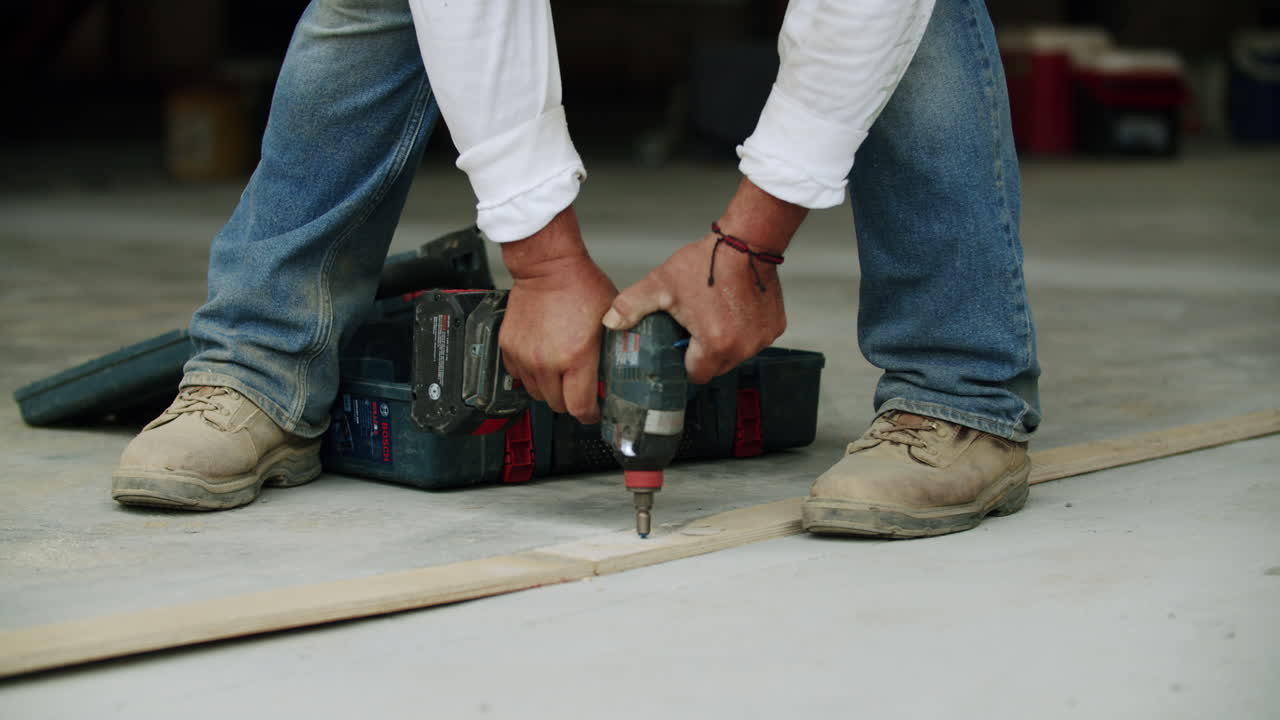 A person using a power drill to secure a screw into a wooden plank on the ground