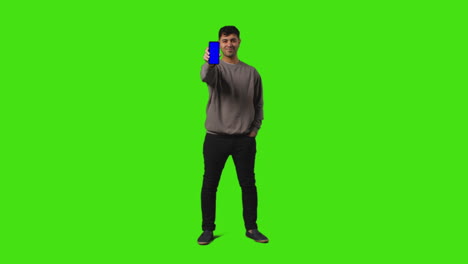 Full-Length-Shot-Of-Man-Holding-Up-Blue-Screen-Mobile-Phone-To-Camera-Against-Green-Screen-Background