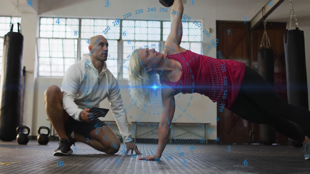 Coach tapping tablet as woman trainee rotating, pulling, pressing kettlebell, HUD tracking fitness