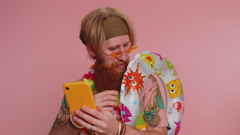 man with a red beard is wearing sunglasses and a hawaiian shirt. he is holding a phone and an inflatable ring. he is smiling and looks happy.