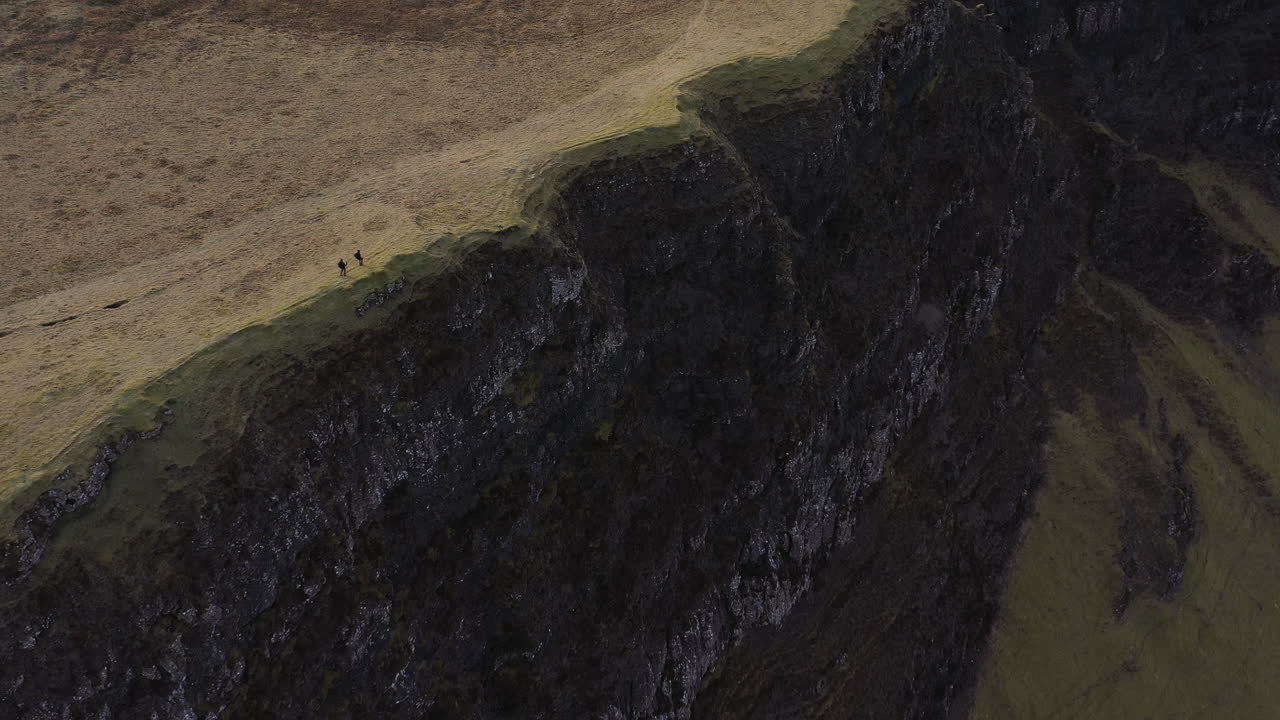 Drone pullback showing two hikers walking along a steep, barren cliff edge on the Trotternish Ridge, Isle of Skye, with exposed erosion and glacially carved landscape.