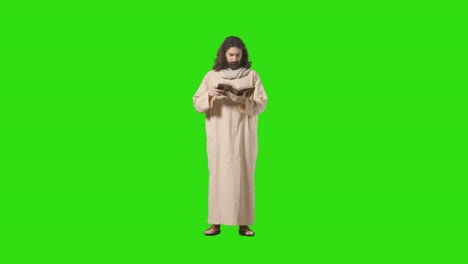 studio shot of man wearing robes and sandals with long hair and beard representing figure of jesus christ reading bible on green screen