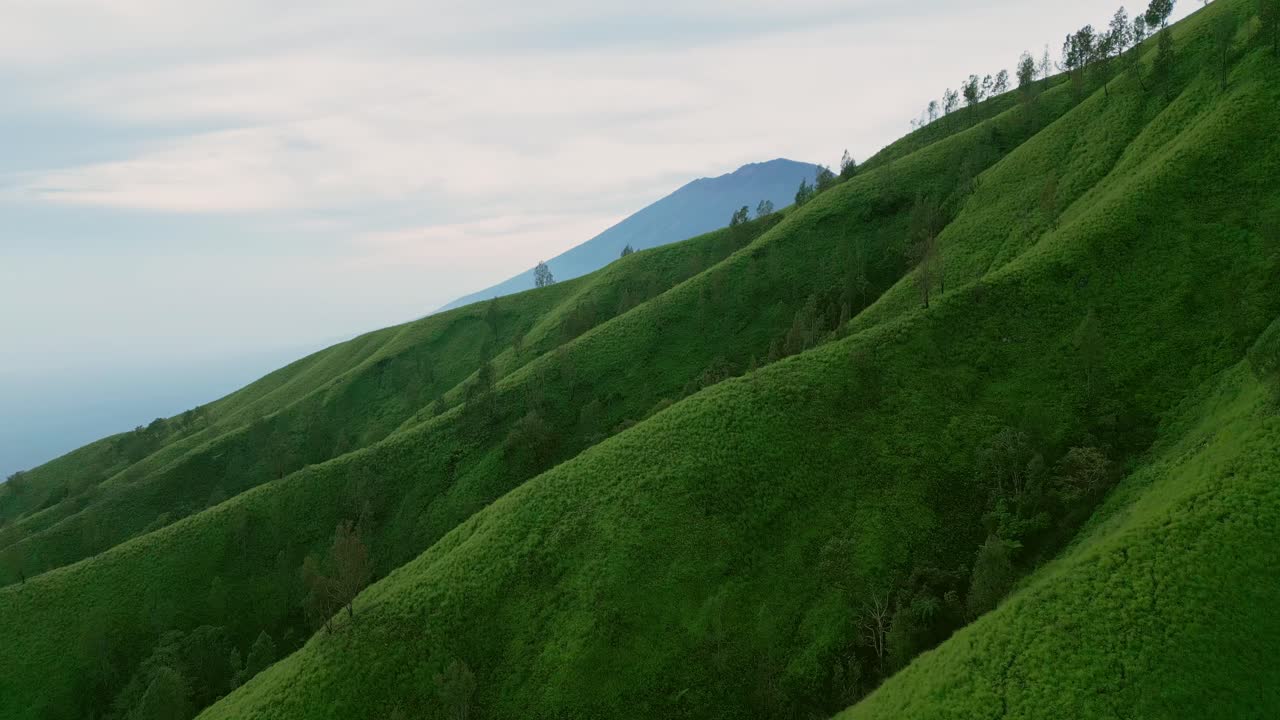 Drone footage reveals the rolling green hills of Trunyan at sunrise as golden light spreads across the landscape with Mount Agung standing tall in the background under a clear tropical sky.