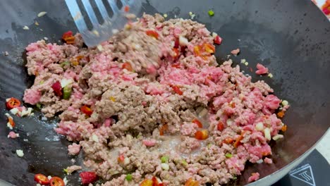 minced meat is cooked with chopped chili peppers in a wok under bright lighting, using a metal spatula to stir and break up the meat