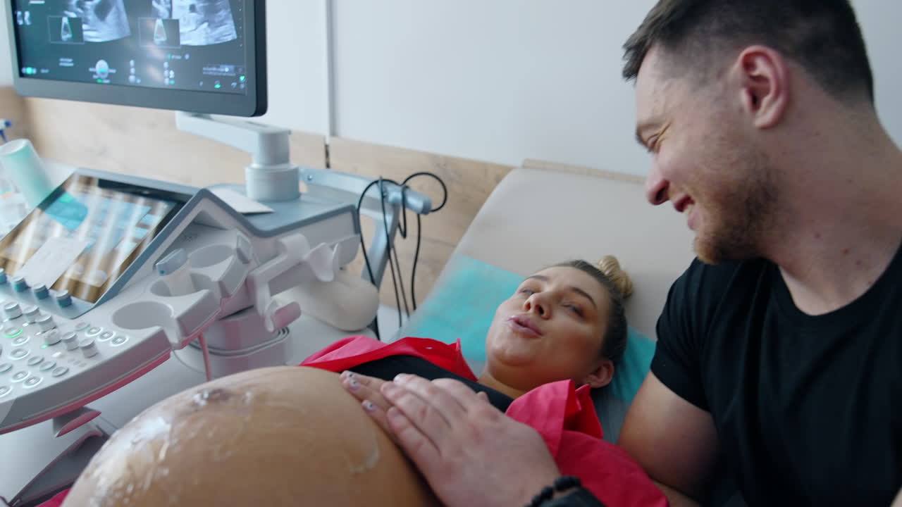 Loving pregnant couple is at doctor's cabinet having ultrasound check up. Man stoking his wife's hand and smiles to her.