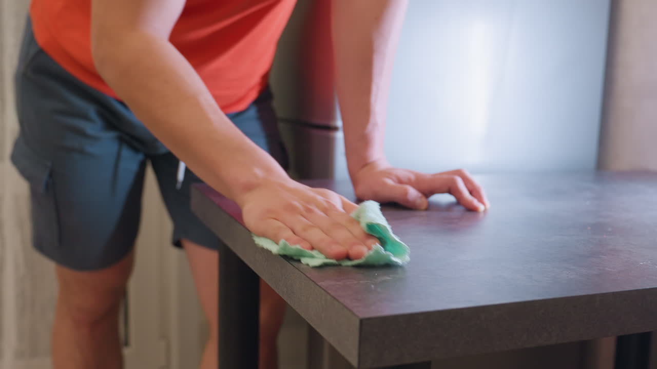 Adult in orange shirt wiping dark kitchen table surface with green cleaning cloth, focusing on removing dust, stains, and dirt, demonstrating household hygiene
