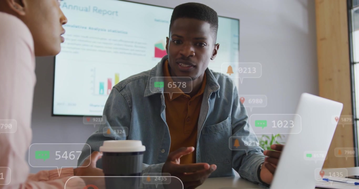 Woman speaking initiating overlaying data bubbles as man pointing at laptop reviewing annual charts